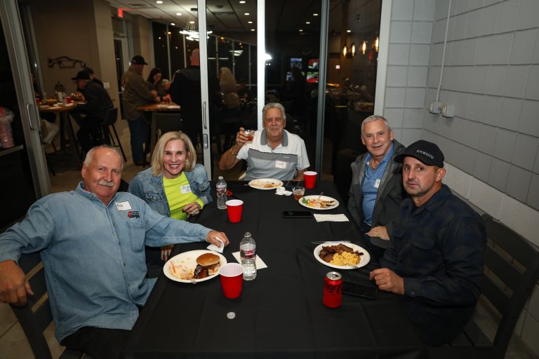 Five adults seated around a black-clothed table at a dining event, smiling for the camera with plates, cups, and drinks in front of them.