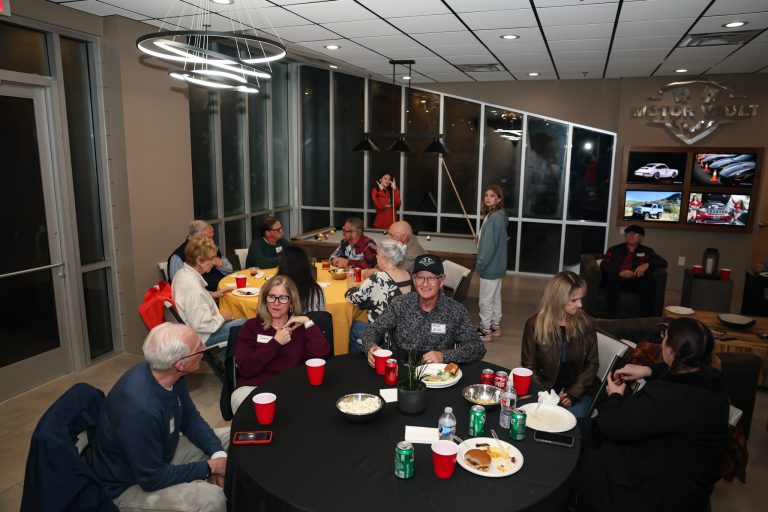 Group of people seated around round tables with plates and red cups at a casual party in a showroom setting.