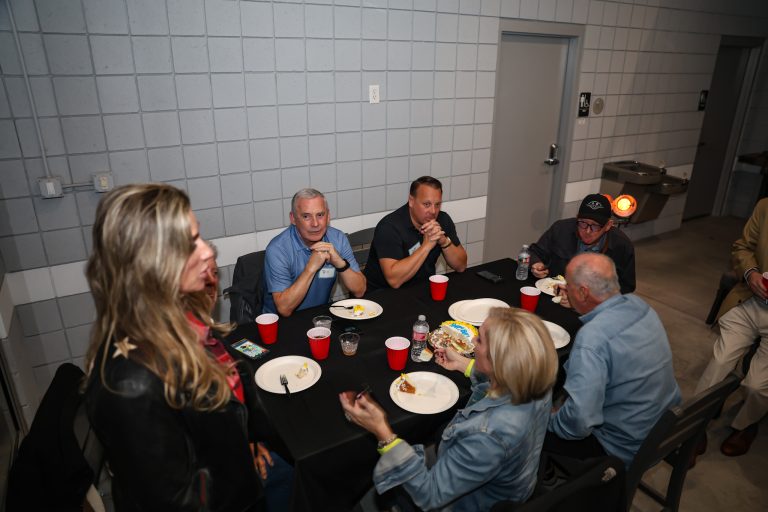 Group of adults seated around a long table eating cake at a casual gathering in a gray-tiled room.