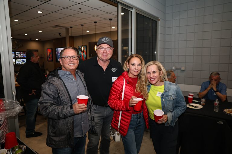 Group of four adults posing for a photo at a casual indoor gathering, holding red cups and smiling at the camera (name tags visible).