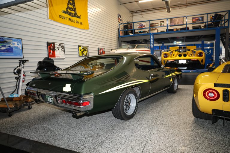 Indoor showroom garage with classic green muscle car in foreground and a bright yellow supercar in the background on a lift, flags and posters on the walls.