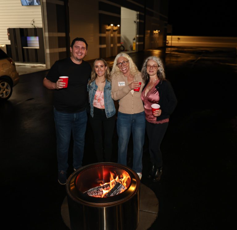 Four adults posing together by a lit outdoor fire pit at night, smiling for the camera.