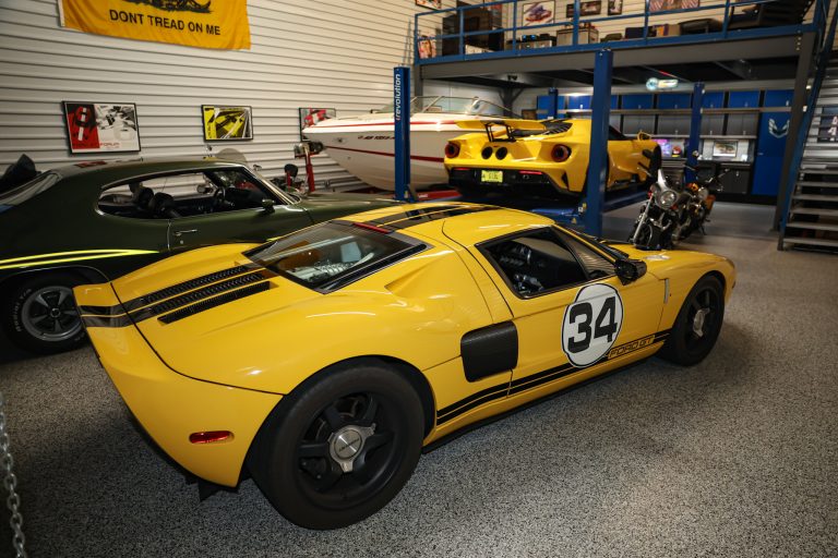 Yellow race car with number 34 in the foreground inside a showroom, a yellow car on a lift behind it, and a green car to the left.