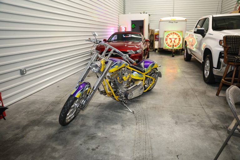 Colorful custom motorcycle with flame paint in the foreground inside a garage, with a red car and neon signs in the background.