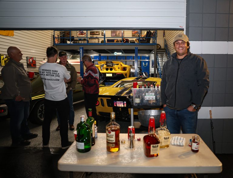 Group of men chatting in a car workshop next to a yellow sports car, with bottles and cups on a table in the foreground and an upper mezzanine in the background.