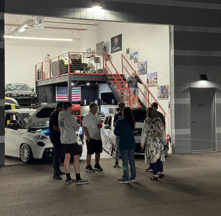 Group of people standing and talking in a car showroom with several cars on display and an upstairs loft area with a red staircase and an American flag in the background.
