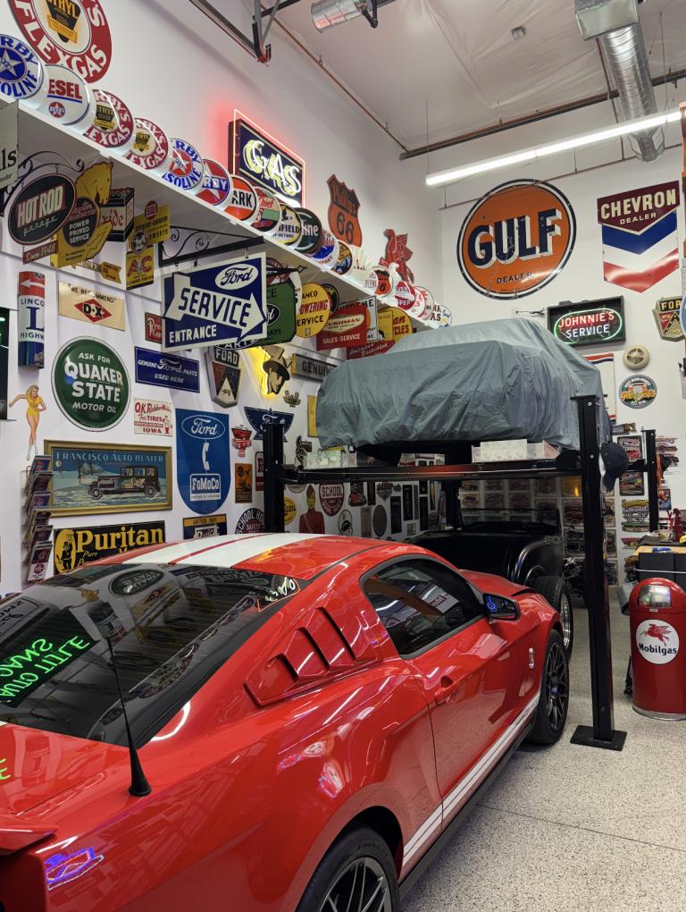 Red sports car in a vintage-sign filled garage, with a car lift and a covered vehicle in the background.