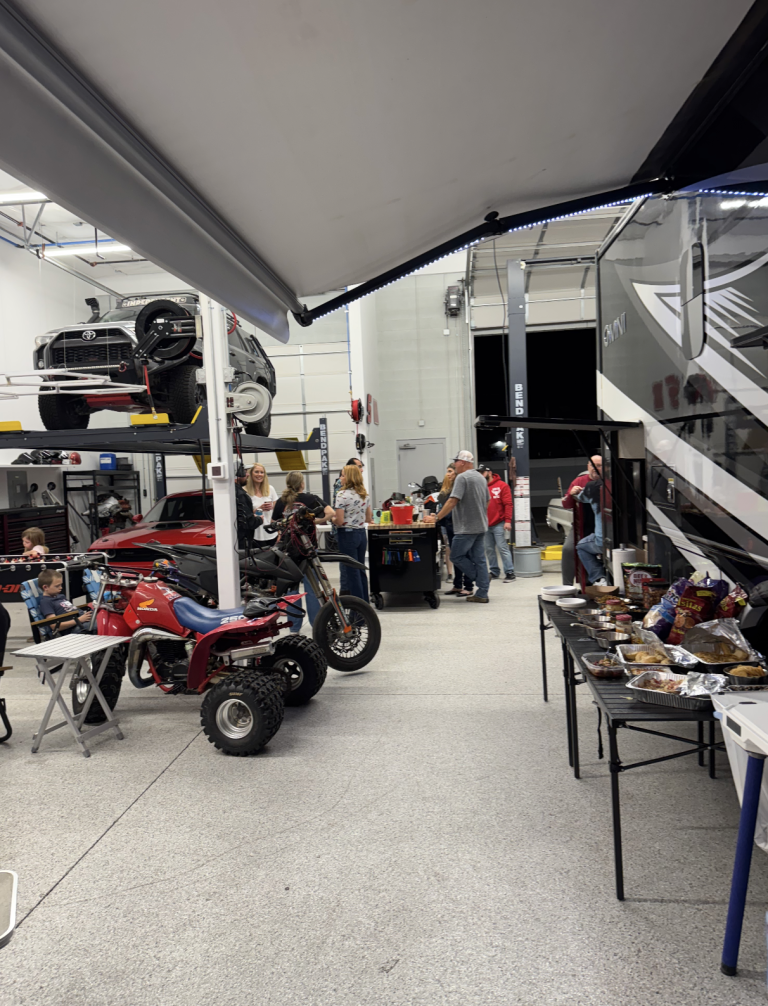 People gathering in a large automotive workshop with vehicles on lifts and a snack table to the side.