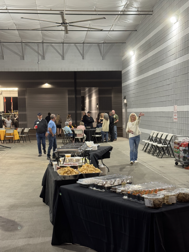Buffet-style tables with snacks in the foreground as groups of people mingle in a large indoor event space; a woman in a light sweater waves toward the camera.