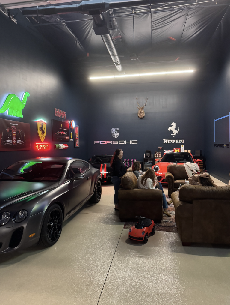 Indoor luxury car showroom with a silver sports car in the foreground, neon Ferrari sign, and a seating area with people nearby.