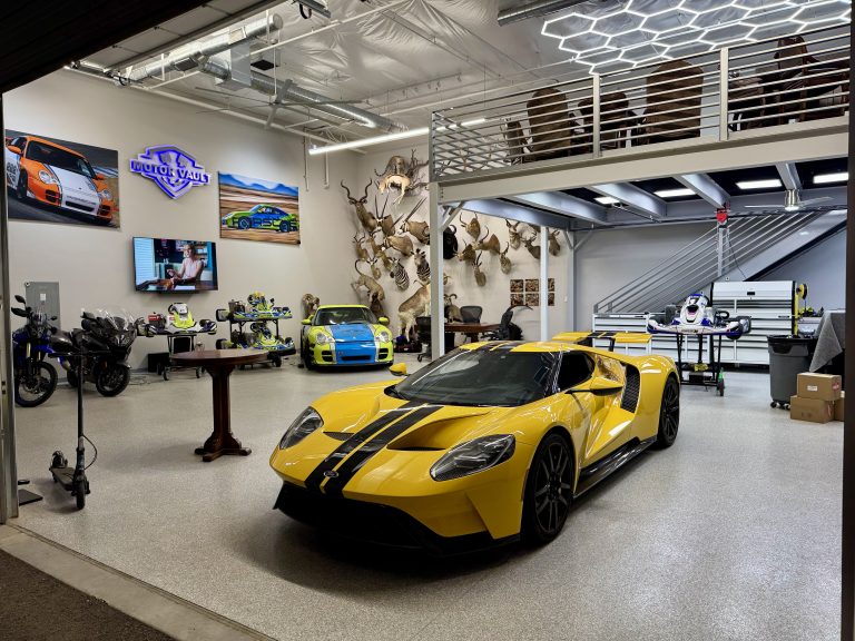 Interior of an automotive showroom featuring bright yellow sports cars, go-karts, and motorcycles; wall of mounted animal trophies and mezzanine seating above.